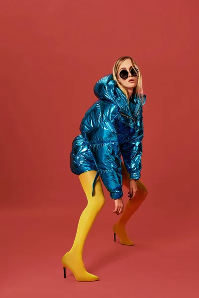 Atractivo flaco joven rubia posando en el estudio sobre fondo rojo. Ella está usando chaqueta azul, gafas de sol, medias amarillas — Foto de Stock