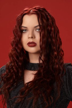 Güzel seksi kadın kırmızı bir arka plan üzerine örme elbiseyle poz. Akşam makyaj. Uzun, sağlıklı, kırmızı kıvırcık saç.