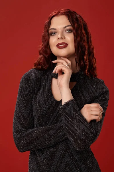 Beautiful sexy woman is posing in a knitted dress on a red background. Evening makeup. Long, healthy, red curly hair.