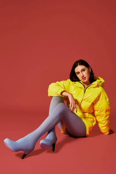 Retrato de estudio de una joven morena sentada en el suelo con chaqueta amarilla y pantalón azul gris mangueras o medias — Foto de Stock