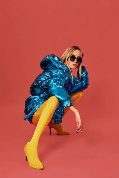 Tineri încrezător blondă fată în jachetă albastră squats în studio înainte de aparat de fotografiat, fundal roșu — Fotografie, imagine de stoc
