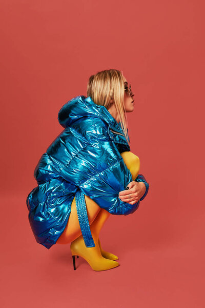 Young cute nice blonde girl in blue jacket sits in a fetal position in studio before camera, red background. Side view