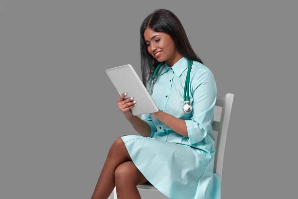 Adorable indian female doctor nurse with stethoscope in aquamarine dress sitting on chair on grey background