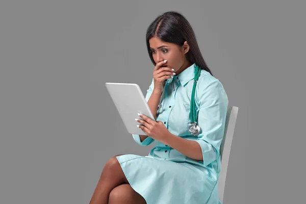 Aranyos indiai női orvos ápolónő sztetoszkóp akvamarin ruha ül a székre, a szürke háttér — Stock Fotó