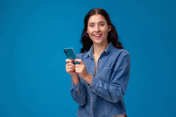 Jovem morena está posando com um smartphone em pé no fundo azul . — Fotografia de Stock