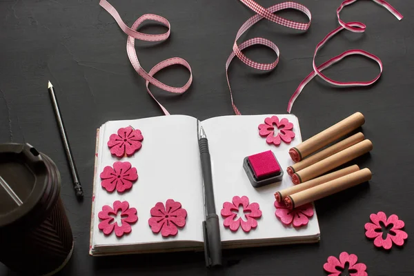 Top view of an empty notebook, scrapbook accessories and a cup of coffee on a black background.