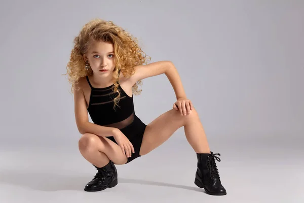 Beautiful little girl gymnast in a black sports swimsuit and boots on a gray background.