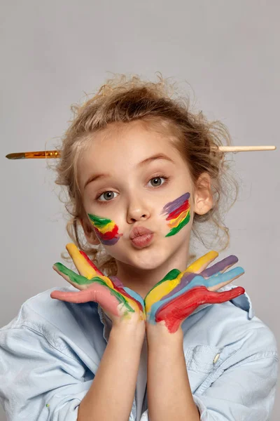 Beautiful little girl with a painted hands and cheeks is posing on a gray background. — Stock Photo, Image