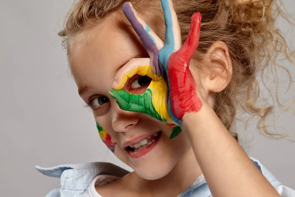 Mooi klein meisje met een geschilderde handen en wangen poseren op een grijze achtergrond. — Stockfoto