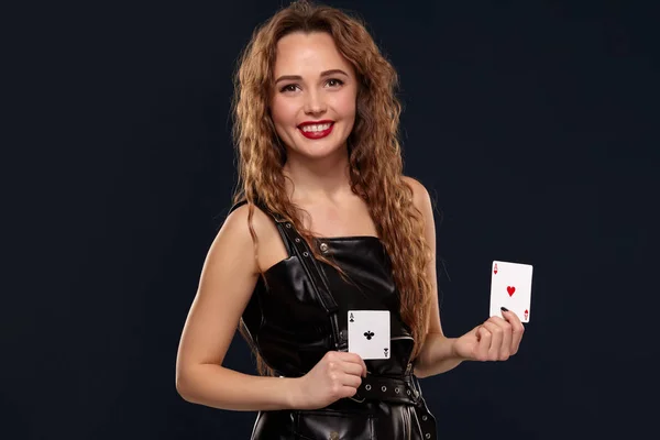 Emotional pretty young redhead woman holding pair of aces in black leather dress on black background — Stock Photo, Image