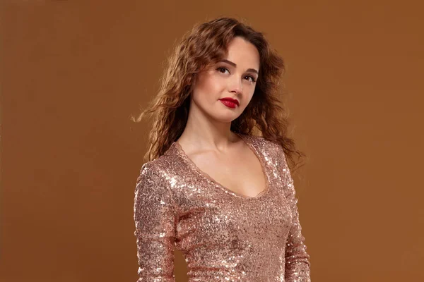 Portrait of young caucasian female in golden cocktail dress looking into the camera on brown background — Stock Photo, Image