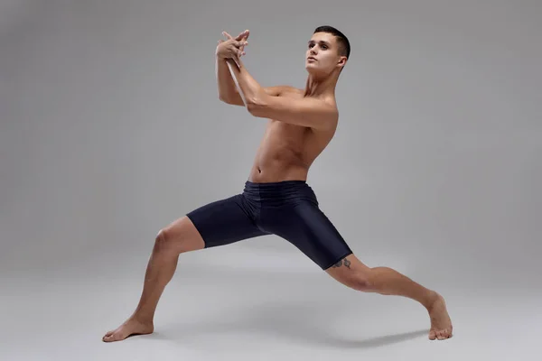 Foto de un hombre guapo bailarín de ballet, vestido con pantalones cortos negros, haciendo un elemento de baile sobre un fondo gris en el estudio . —  Fotos de Stock