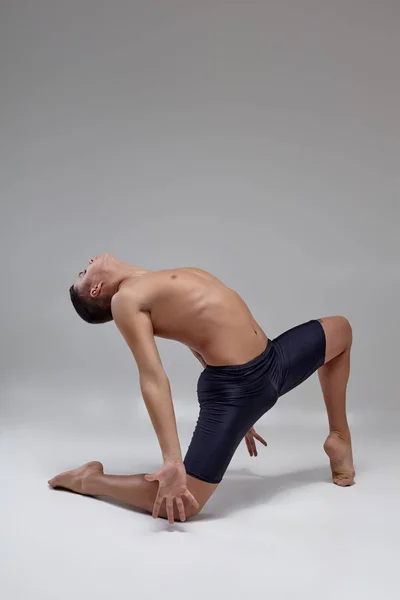 Foto de um homem bonito bailarino de balé, vestido com shorts pretos, fazendo um elemento de dança contra um fundo cinza no estúdio . — Fotografia de Stock