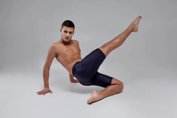 Foto de um homem bonito bailarino de balé, vestido com shorts pretos, fazendo um elemento de dança contra um fundo cinza no estúdio . — Fotografia de Stock