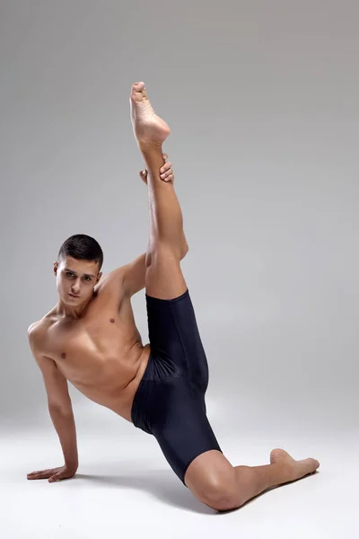Foto de un hombre guapo bailarín de ballet, vestido con pantalones cortos negros, haciendo un elemento de baile sobre un fondo gris en el estudio . —  Fotos de Stock