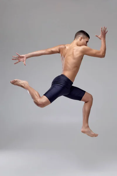 Foto de um homem bonito bailarino de balé, vestido com shorts pretos, fazendo um elemento de dança contra um fundo cinza no estúdio . — Fotografia de Stock