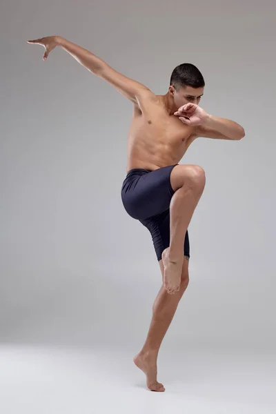 Foto de um homem bonito bailarino de balé, vestido com shorts pretos, fazendo um elemento de dança contra um fundo cinza no estúdio . — Fotografia de Stock