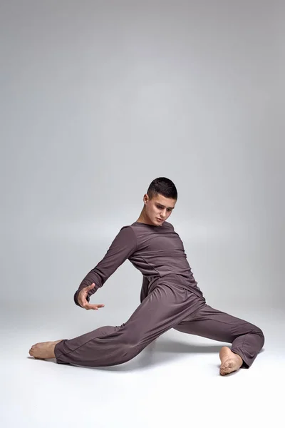 Photo d'un danseur de ballet homme athlétique vêtu d'un survêtement gris, faisant un élément de danse sur un fond gris en studio . — Photo