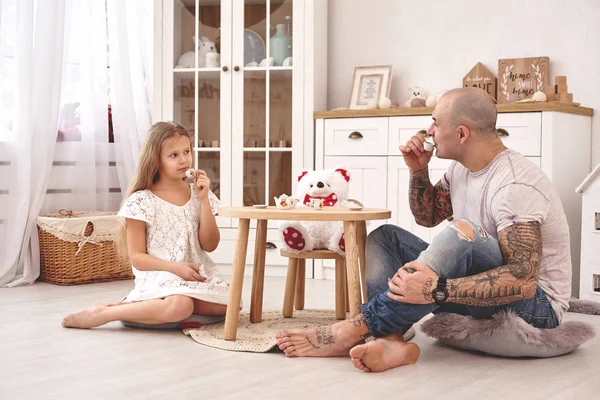 Adorable daughter wearing a white dress whith her loving father. They are drinking tea from a toy dishes in a modern kids room. Happy family.