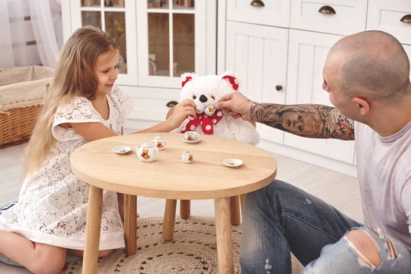 Rozkošná dcera nosí v bílých šatech svůj milující otec. Popíjí čaj z hračkářská jídla v moderním dětovém pokoji. Šťastná rodina. — Stock fotografie