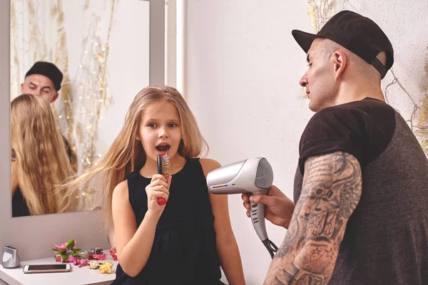 Roztomilá dceruška a její Tetovaný otec si hrají vedle zrcadla. Táta dělá své dcery vlasy. Rodinné svátky. — Stock fotografie