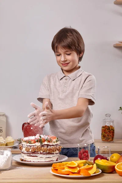 Kisfiú, hogy egy házi készítésű sütemény egy könnyű recept a konyhában szemben egy fehér falat polcok rajta. — Stock Fotó