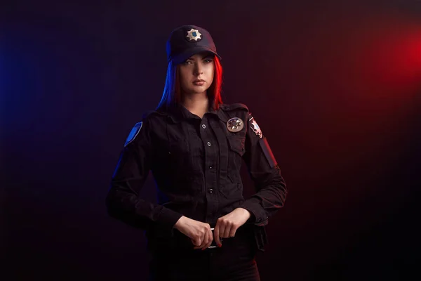 Sério policial feminino está posando para a câmera contra um fundo preto com backlighting vermelho e azul . — Fotografia de Stock