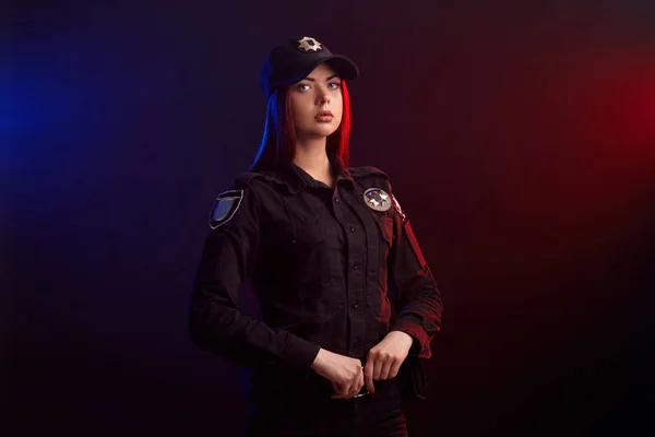 Une policière sérieuse pose pour la caméra sur un fond noir avec un rétroéclairage rouge et bleu . — Photo