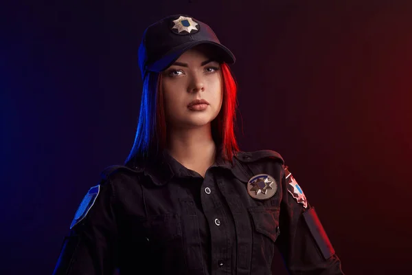 Nahaufnahme einer seriösen Polizistin, die vor schwarzem Hintergrund mit roter und blauer Hintergrundbeleuchtung für die Kamera posiert. — Stockfoto