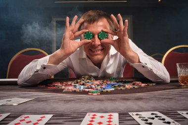 Handsome emotional man is playing poker sitting at the table in casino. clipart