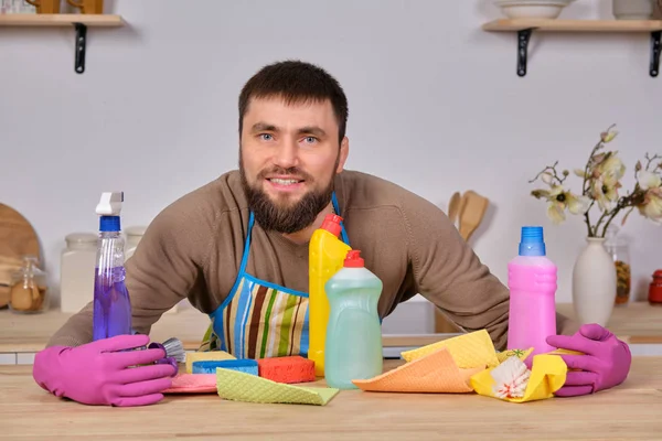 Jonge knappe bebaarde man in de keuken, toont al zijn schoonmaakpersoneel - wasmiddelen, borstels, sprays. Hij denkt dat hij klaar is voor echte schoonmaak. — Stockfoto
