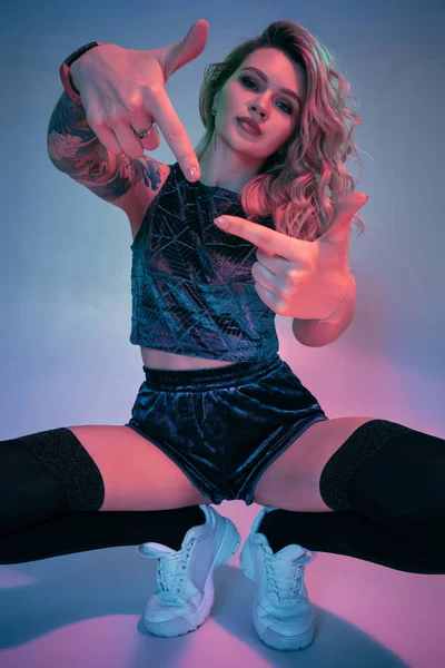 Young fitness blonde woman wearing blue velour booty shorts squats in front of camera, looking at camera, pink and blue background — 스톡 사진