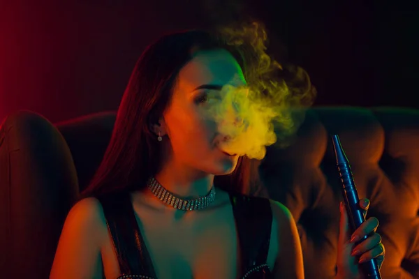 Sexy morena modelo é fumar um narguilé exalando uma fumaça em um clube noturno de luxo . — Fotografia de Stock