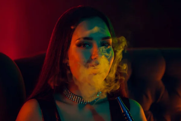 Sexy morena modelo é fumar um narguilé exalando uma fumaça em um clube noturno de luxo . — Fotografia de Stock