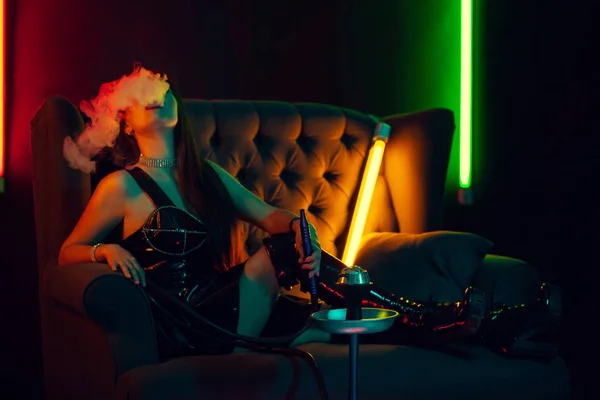 Sexy morena modelo é fumar um narguilé exalando uma fumaça em um clube noturno de luxo . — Fotografia de Stock