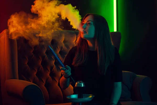Красивая девушка в черной футболке курит кальян, выдыхая сигарету в роскошном ночном клубе . — стоковое фото