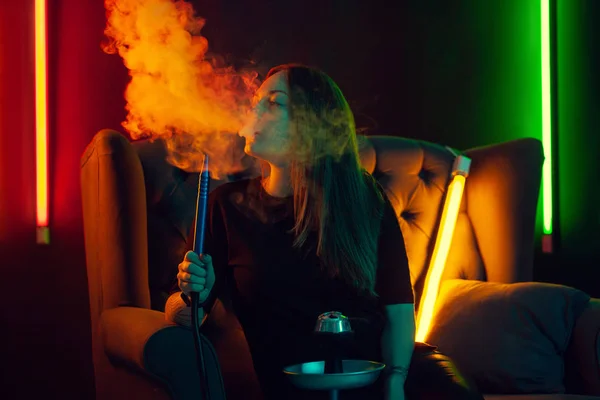 Menina bonita em uma camiseta preta está fumando um narguilé exalando um cigarro em um clube noturno de luxo . — Fotografia de Stock