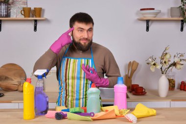 Mutfaktaki yakışıklı sakallı genç adam temizlik malzemelerini gösteriyor. Deterjanlar, fırçalar, spreyler. Gerçek bir temizliğe hazır olduğunu düşünüyor.