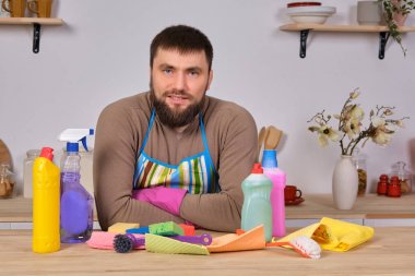 Mutfaktaki yakışıklı sakallı genç adam temizlik malzemelerini gösteriyor. Deterjanlar, fırçalar, spreyler. Gerçek bir temizliğe hazır olduğunu düşünüyor.