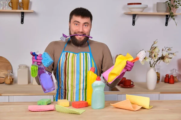Confused young handsome bearded man in the kitchen, have no idea what to do with all his detergents, brushes, sprays