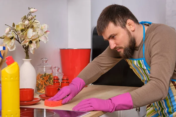 Giovane uomo barbuto bello in cucina, indossando grembiule e guanti rosa pulisce la superficie di cottura con detergenti — Foto Stock