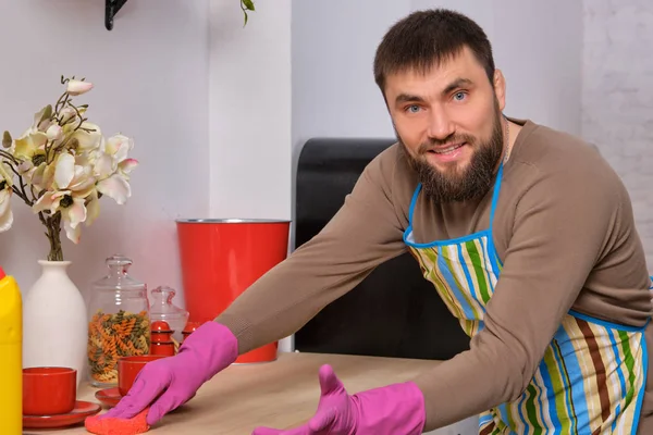 Homem barbudo bonito jovem na cozinha, tentando limpar uma mesa usando detergentes, escovas, sprays — Fotografia de Stock