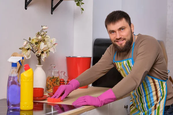 Joven hombre barbudo guapo en la cocina, con delantal y guantes de color rosa limpia la superficie de cocción con detergentes — Foto de Stock