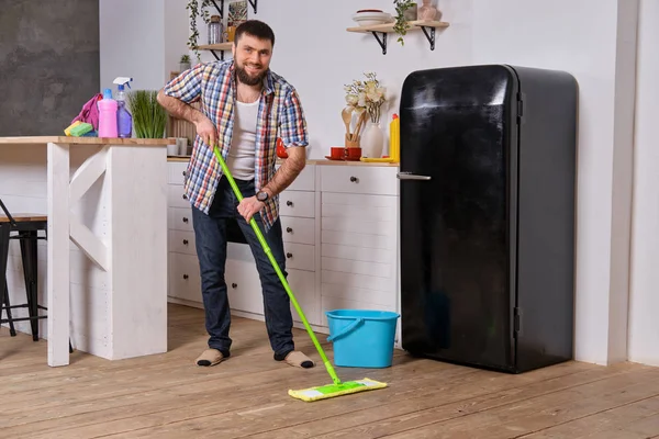 El ama de llaves del marido limpia la cocina. Joven hombre feliz con fregona verde lava el suelo — Foto de Stock