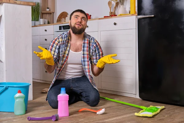 Kaukasische jonge bebaarde man zit op de vloer van zijn keuken en probeert te reinigen met al zijn wasmiddelen, Rags, washandjes en borstels — Stockfoto