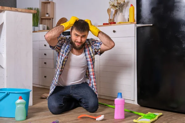 Молодой кавказский бородач сидит на полу своей кухни и пытается почистить ее всеми своими моющими средствами, тряпками, мочалками и щетками. — стоковое фото