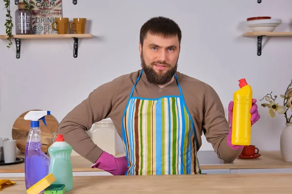 Young bearded man in kitchen makes advertising. He promotes detergent, holding it in one hand and shows at camera — 스톡 사진