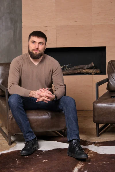 Confident Young handsome serious man sitts on leather chair in living room with hand crossed.