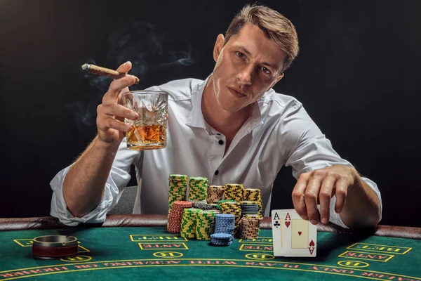 Snygg känslomässig man spelar poker sitter vid bordet i kasino mot en vit spotlight. — Stockfoto