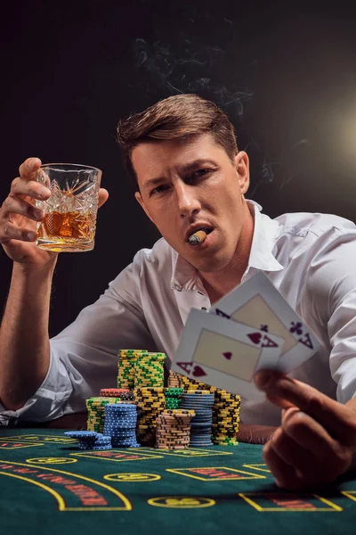 Handsome emotional man is playing poker sitting at the table in casino against a white spotlight.